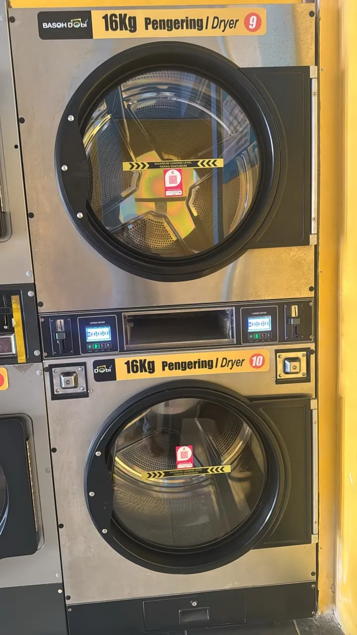 Two stacked 16 kg laundromat dryers with a pink DuitNow QR sticker on each door and the coin slots on the panel between them