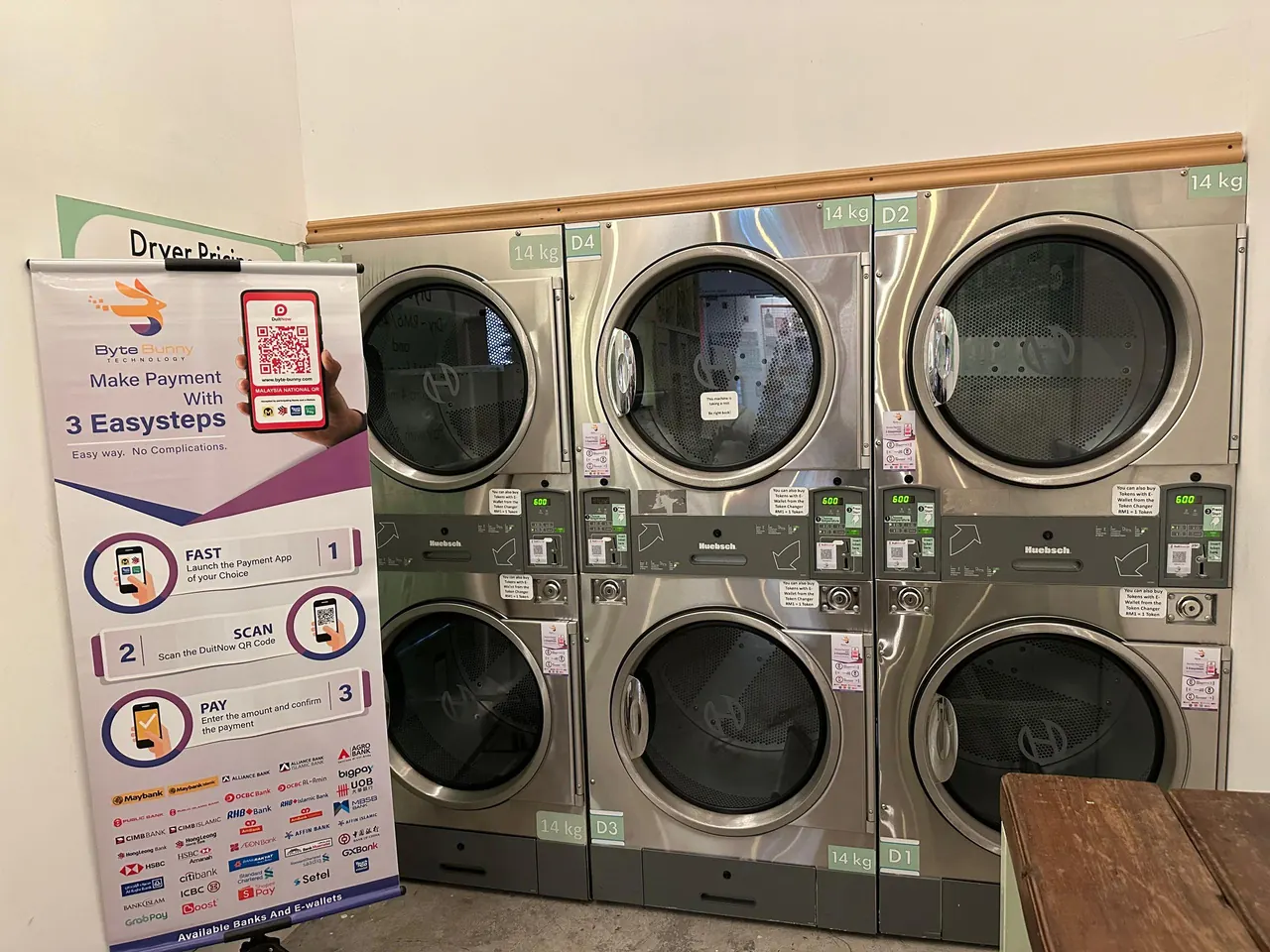 Stacked self-service dryers in a laundromat with a QR sticker on each machine's panel, beside a Byte Bunny standee showing how to pay in three easy steps